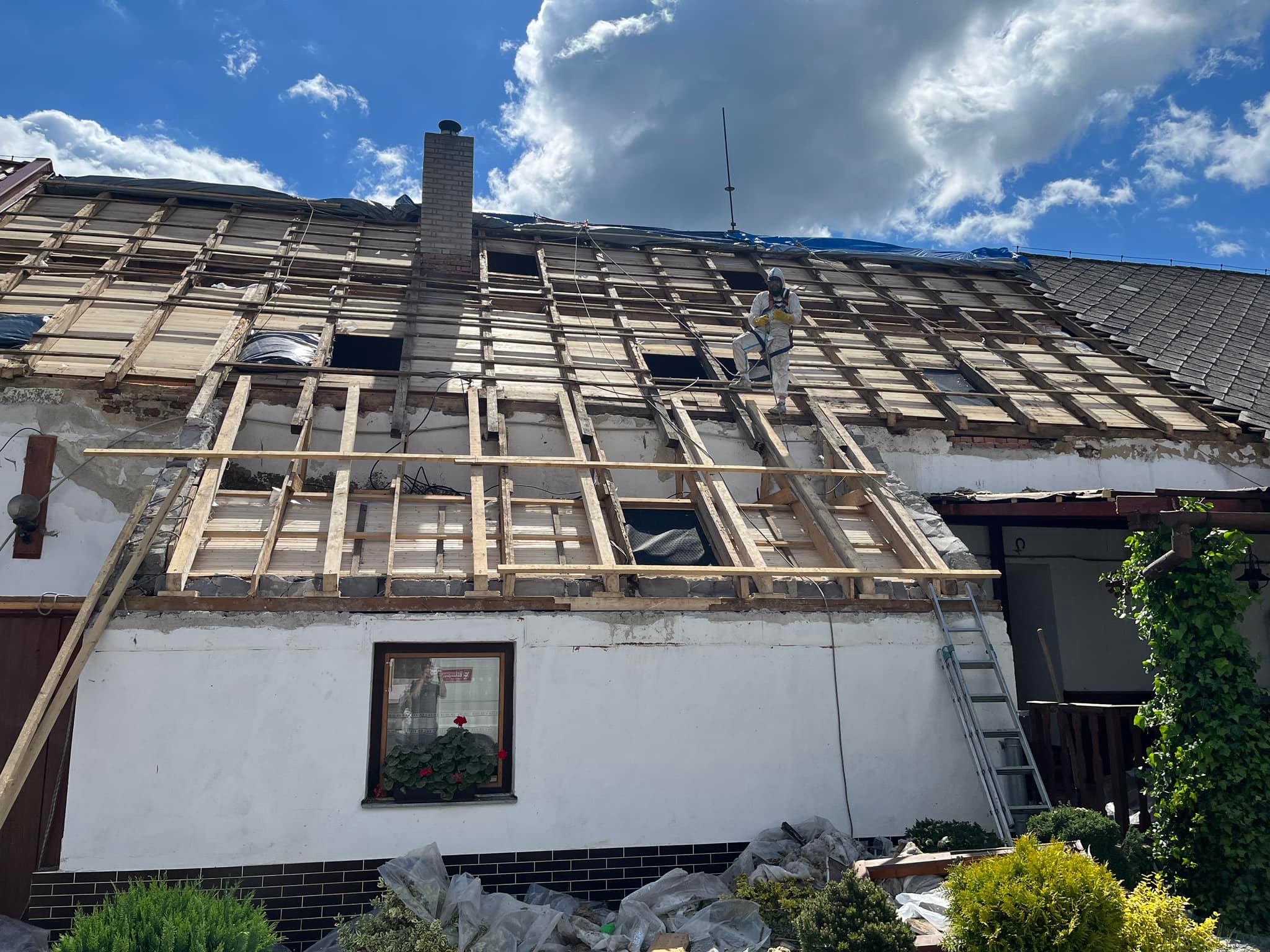 Pracovník v ochranném oblečení opravuje krov rodinného domu, kde jsou části střechy odhalené a viditelné dřevěné trámy.